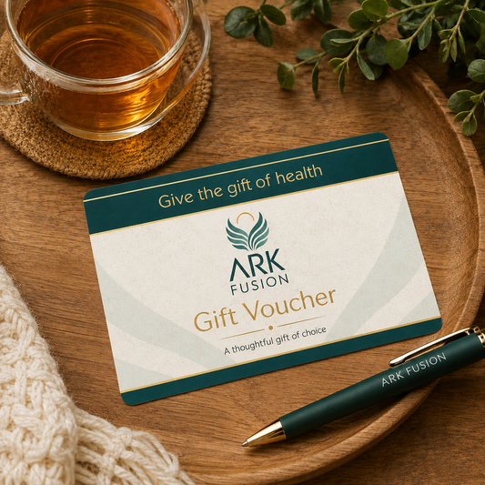 A gift voucher card from ARK FUSION lies on a round wooden tray, styled with a glass cup of tea, a green pen, and a knit sweater. The card has a cream center with dark teal bands and gold text, and the ARK FUSION logo is clearly visible.